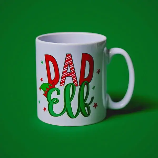 White festive mug with colourful Dad Elf lettering and Christmas-themed decorations displayed against a green background.